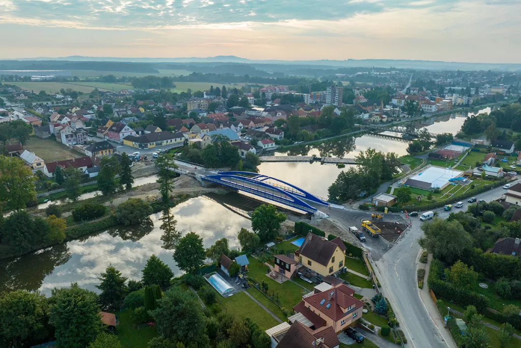 Vue aérienne d’un pont bleu récemment installé au-dessus d’une rivière, au cœur d’une ville résidentielle entourée de verdure et de routes en travaux.